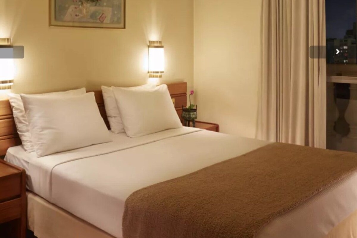 A hotel room with a bed, two bedside tables, a nightstand, a lamp, and a window with curtains.