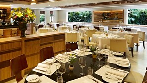 A restaurant with multiple dining tables set for guests, a bar area, and large windows with a view of greenery. - Regent Park Hotel (São Paulo)