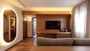 A modern living room with a wooden floor, a large mirror, a flat-screen TV mounted on a wooden panel, and a beige sofa. - Regent Park Hotel (São Paulo)