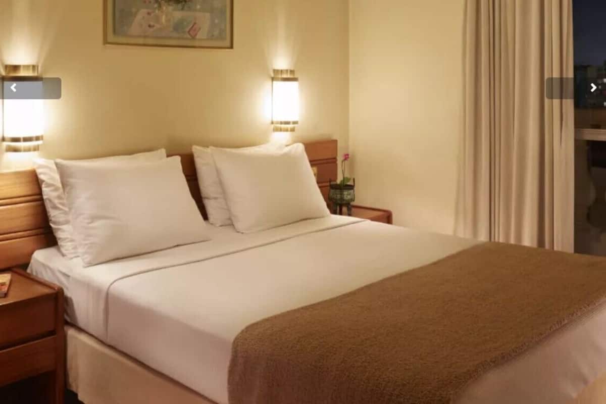 A hotel room with a bed, two bedside tables, a nightstand, a lamp, and a window with curtains.