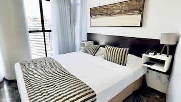 Quarto de hotel moderno com uma cama grande, mesa de cabeceira com abajur, ar condicionado e vista para a cidade pela janela.