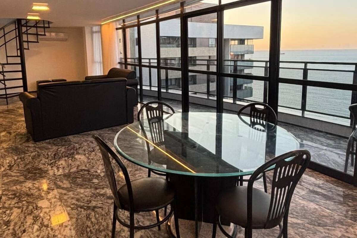 Una sala de estar moderna con una mesa y sillas de vidrio, un sofá y vistas al mar a través de grandes ventanales.