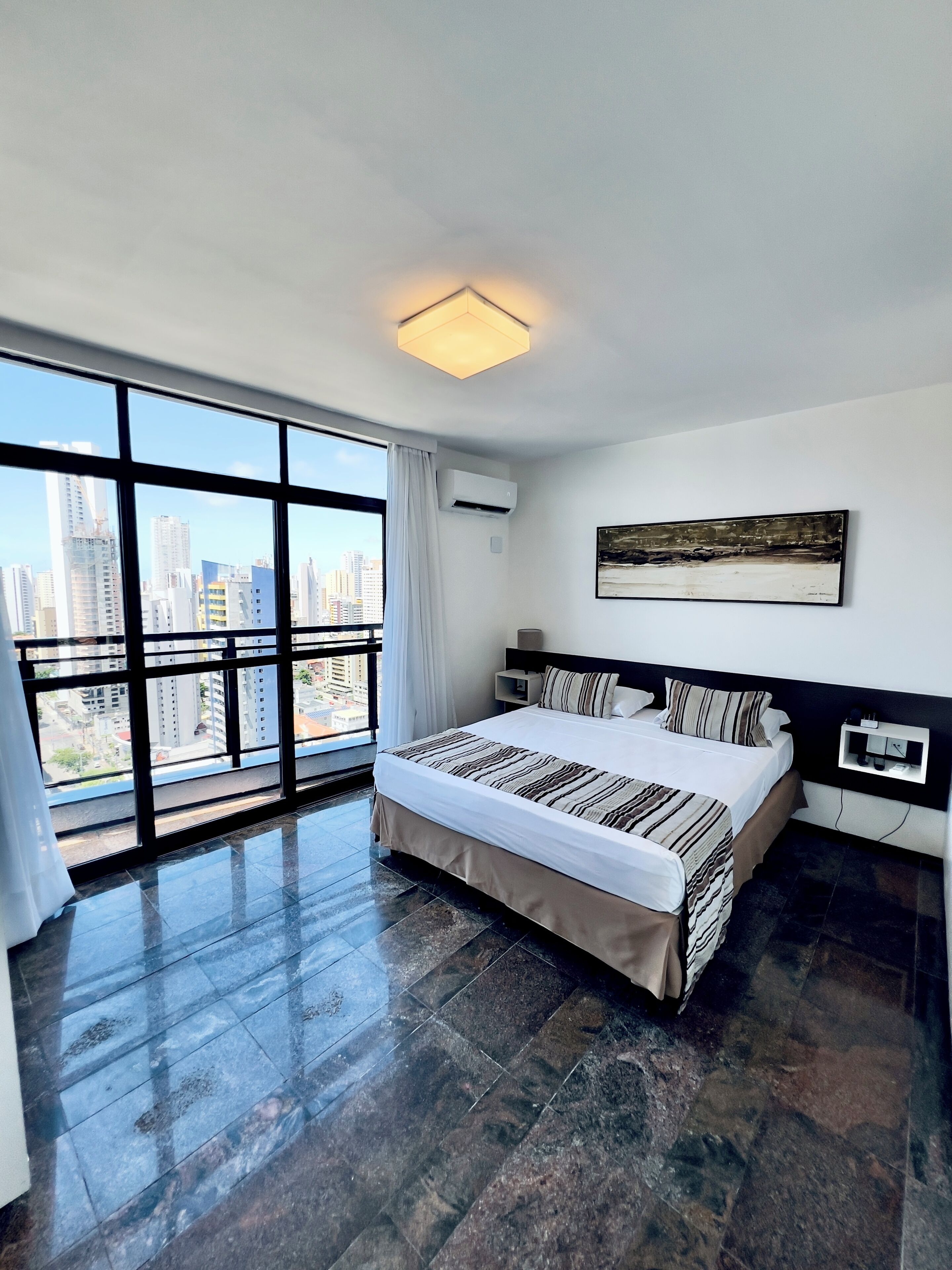 Quarto moderno com uma cama grande, vista para a cidade através de janelas do chão ao teto e ar condicionado fixado na parede.