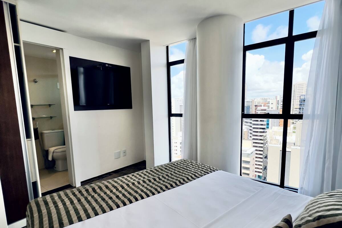 Habitación de hotel con una cama grande, un televisor montado en la pared y vistas de la ciudad a través de amplios ventanales.