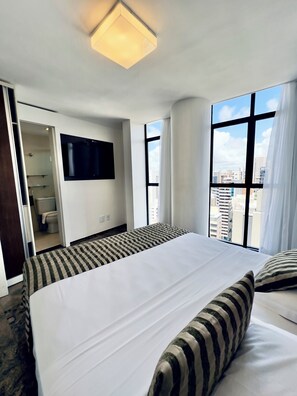 Ein Hotelzimmer mit einem großen Bett, einem an der Wand montierten Fernseher und Blick auf die Stadt durch große Fenster.