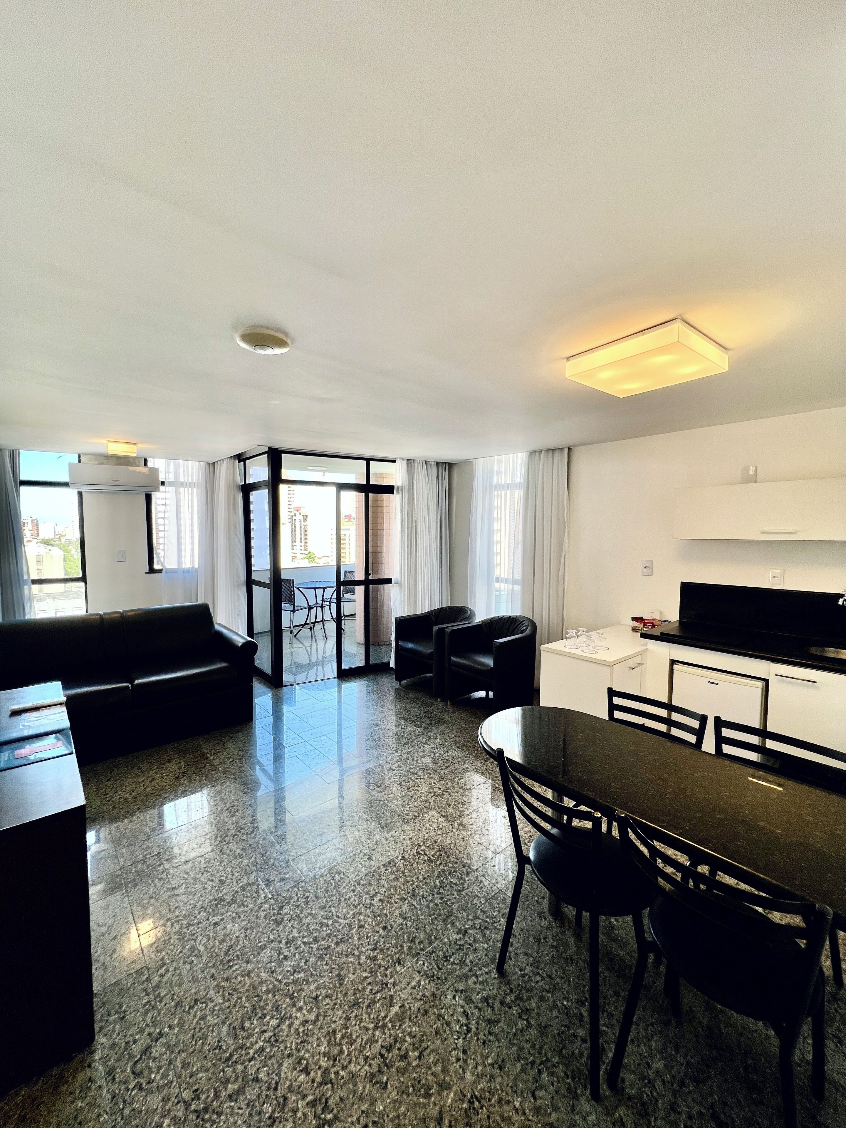 Uma sala de estar moderna e bem iluminada com sofá, mesa de jantar, cadeiras e uma cozinha equipada com frigorífico e micro-ondas.