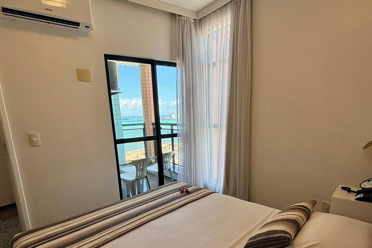 Habitación de hotel con una cama grande, vista al mar y un balcón con sillas.