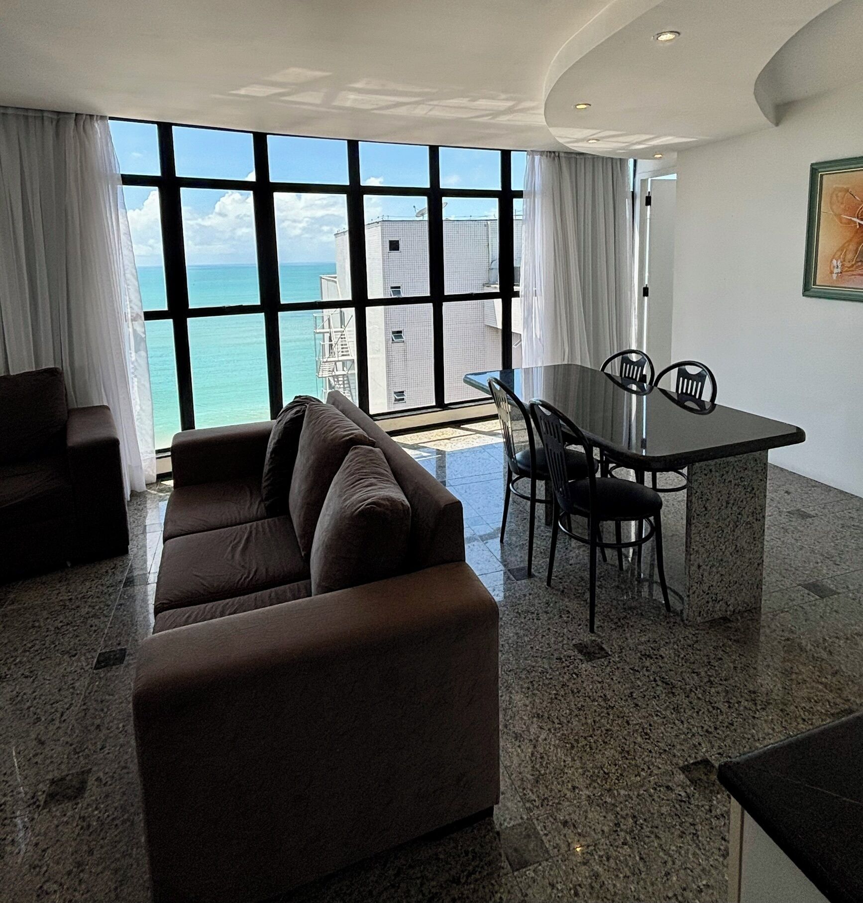 A modern living room with a large window overlooking the ocean, a dark brown sofa, a dining table with chairs, and a framed picture on the wall.