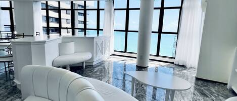 A modern hotel lobby with a marble floor, white sofas, a small round table, and large windows offering a view of the cityscape.