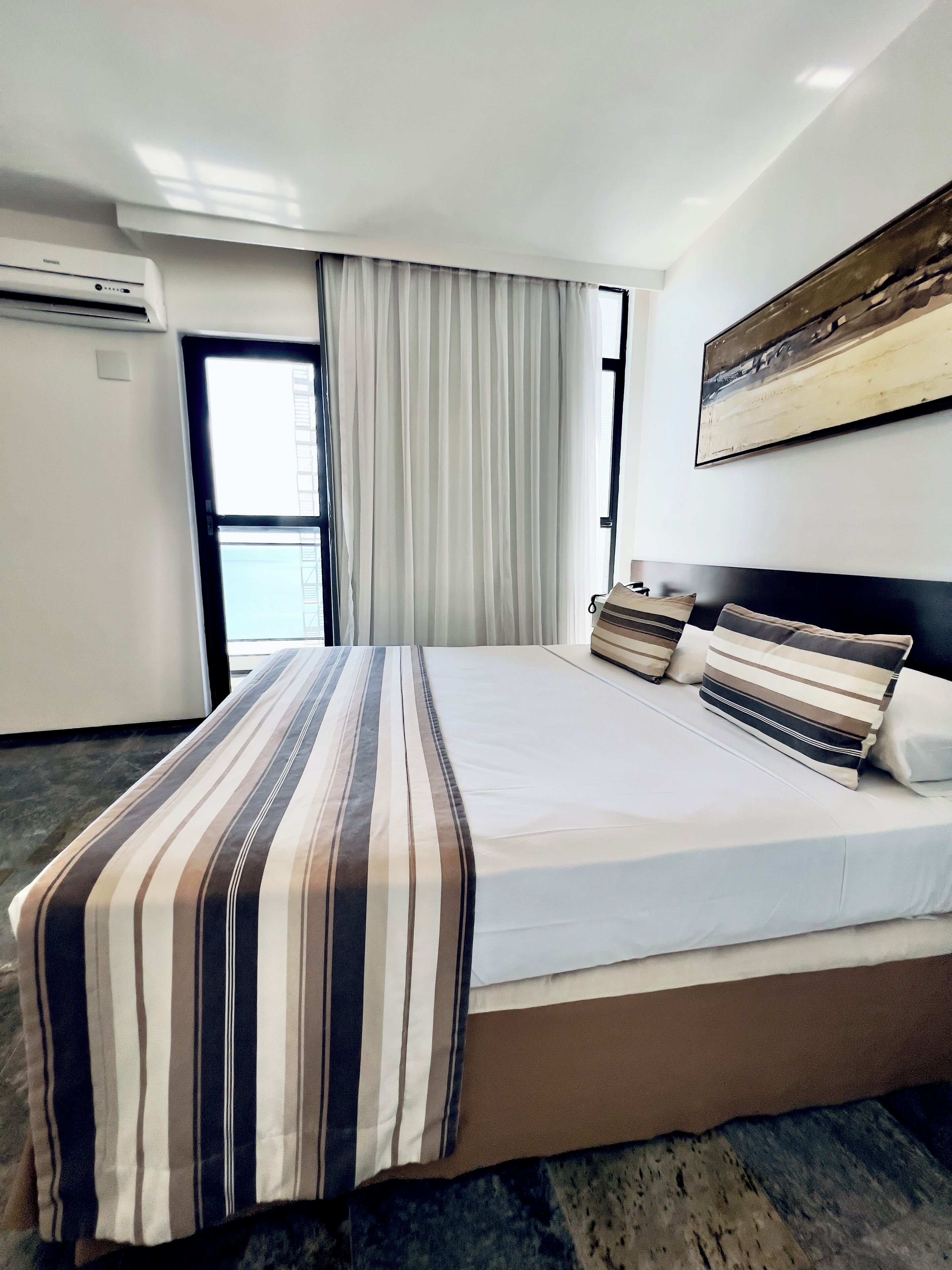 Een hotelkamer met een groot bed, een raam met gordijnen, een airconditioner en een schilderij aan de muur.