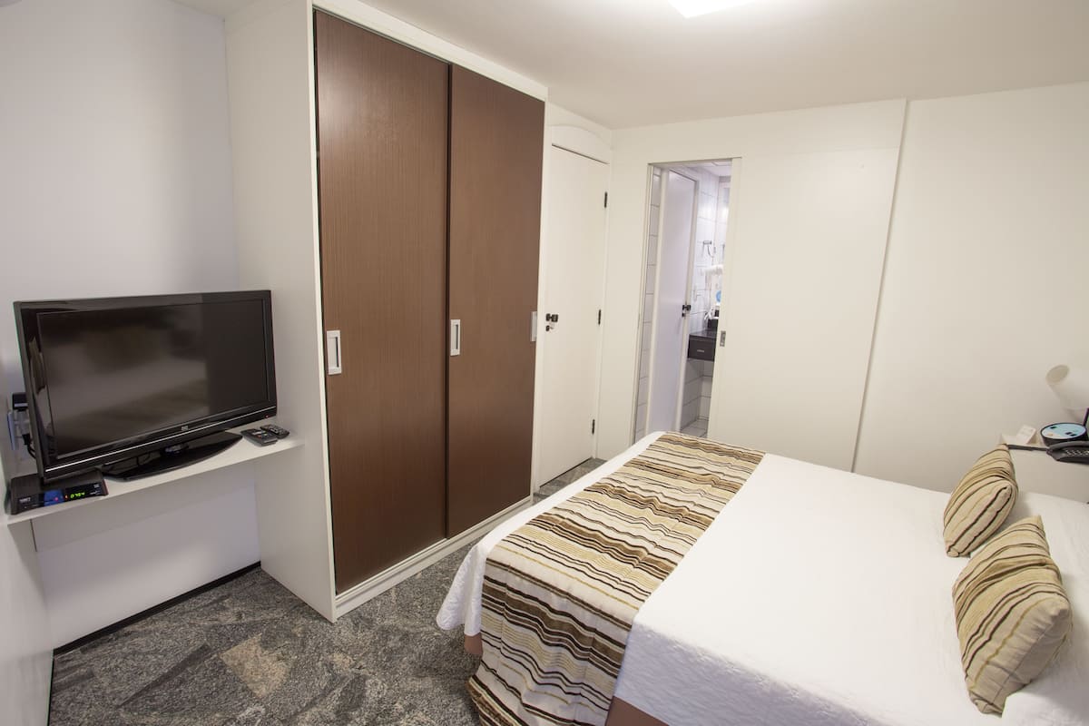 Habitación de hotel con una cama, un televisor sobre un mueble, un armario y un baño visible a través de una puerta abierta.