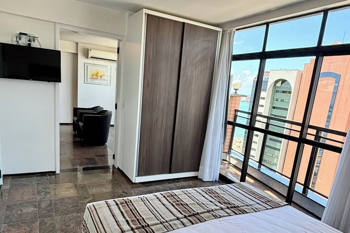 Habitación de hotel con una cama grande, un escritorio, un armario y vistas a la ciudad a través de amplias ventanas.