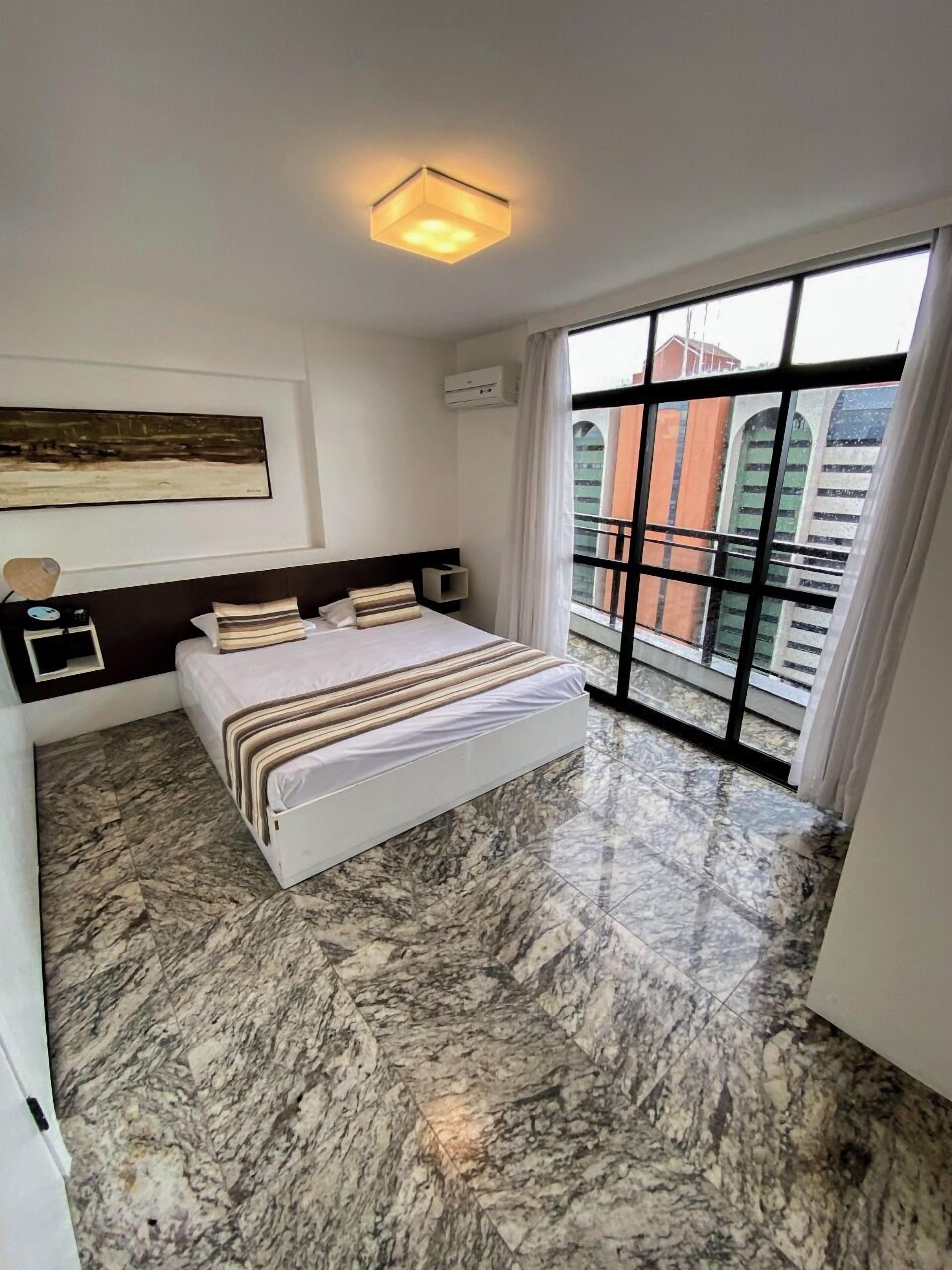 Une chambre moderne avec un grand lit, une tête de lit, un climatiseur fixé au mur, un tableau et une vue sur les bâtiments par une grande fenêtre.
