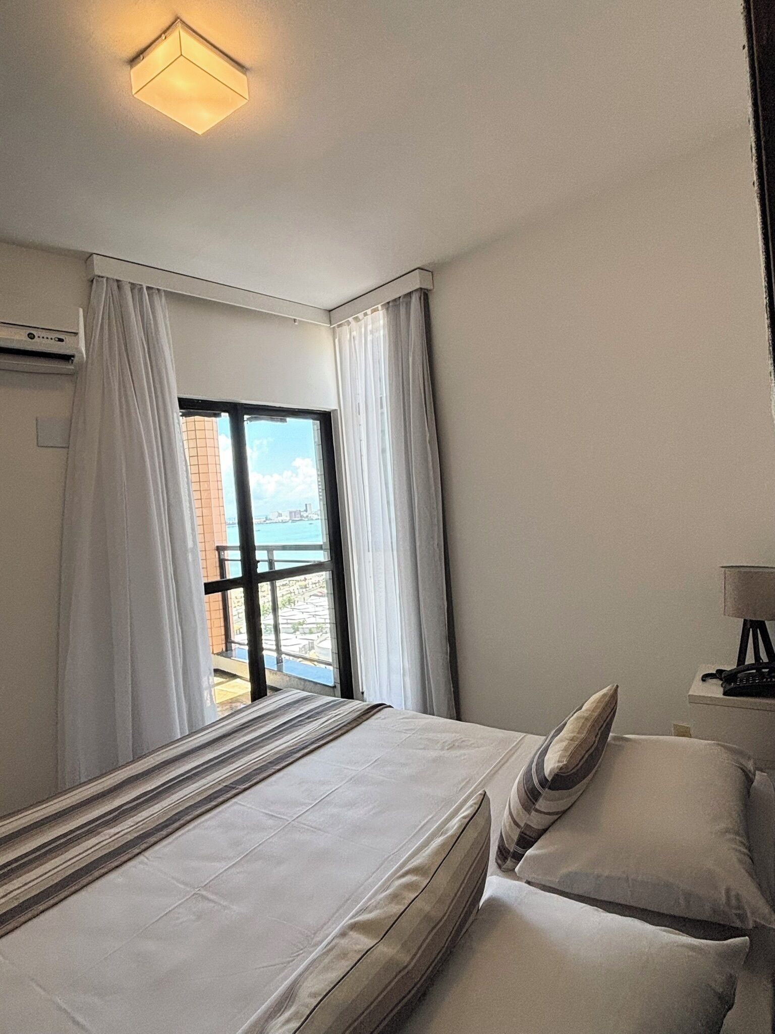 Een hotelkamer met een bed, nachtkastje, lamp en uitzicht op zee door een raam.