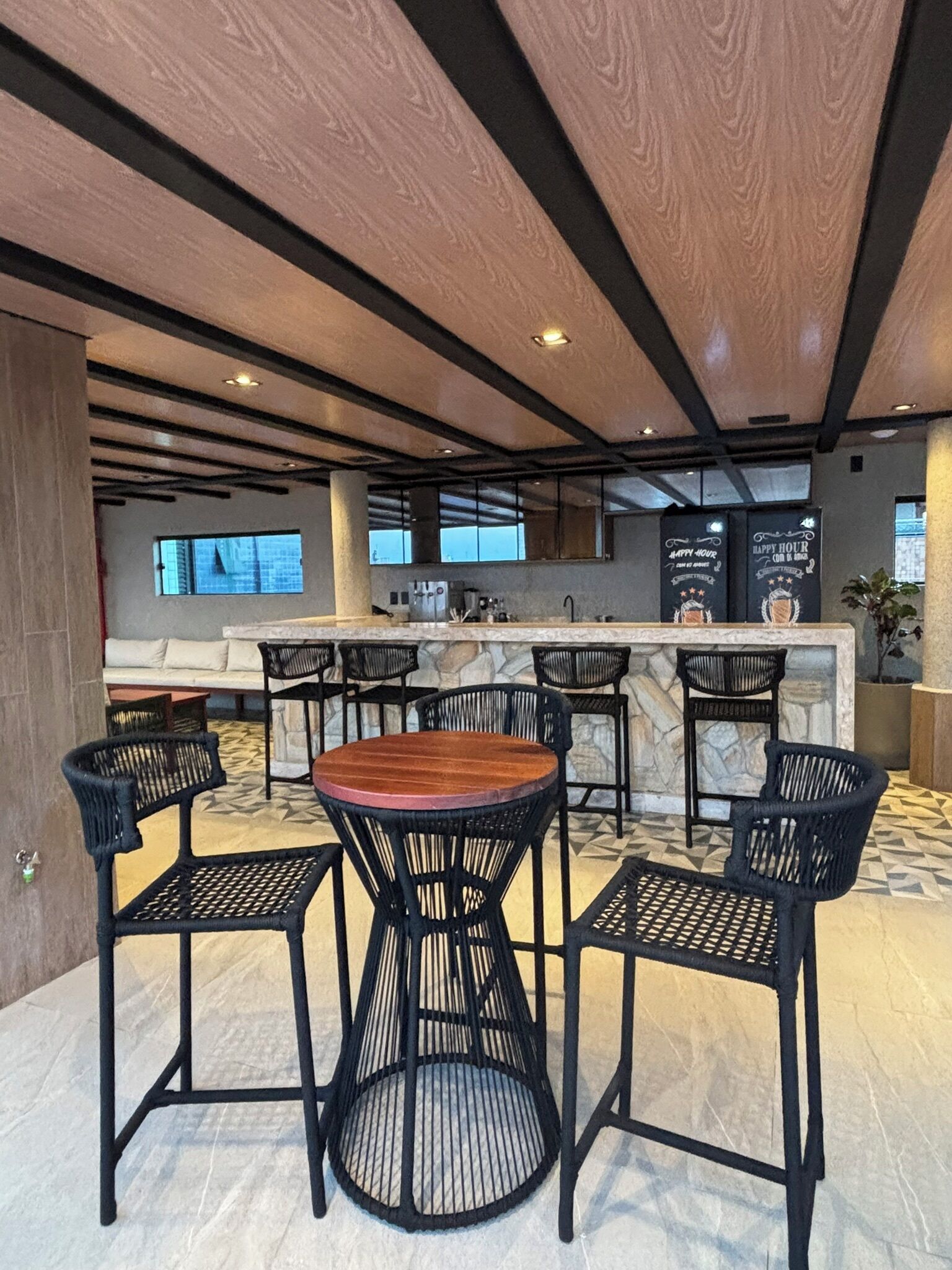 Ein stilvoller Barbereich mit einem runden Tisch in der Mitte und vier schwarzen Stühlen. Die Marmor-Bar verfügt über ein Edelstahl-Spülbecken und eine Kaffeemaschine. Die Decke zeigt freiliegende Balken und eingelassene Beleuchtung.