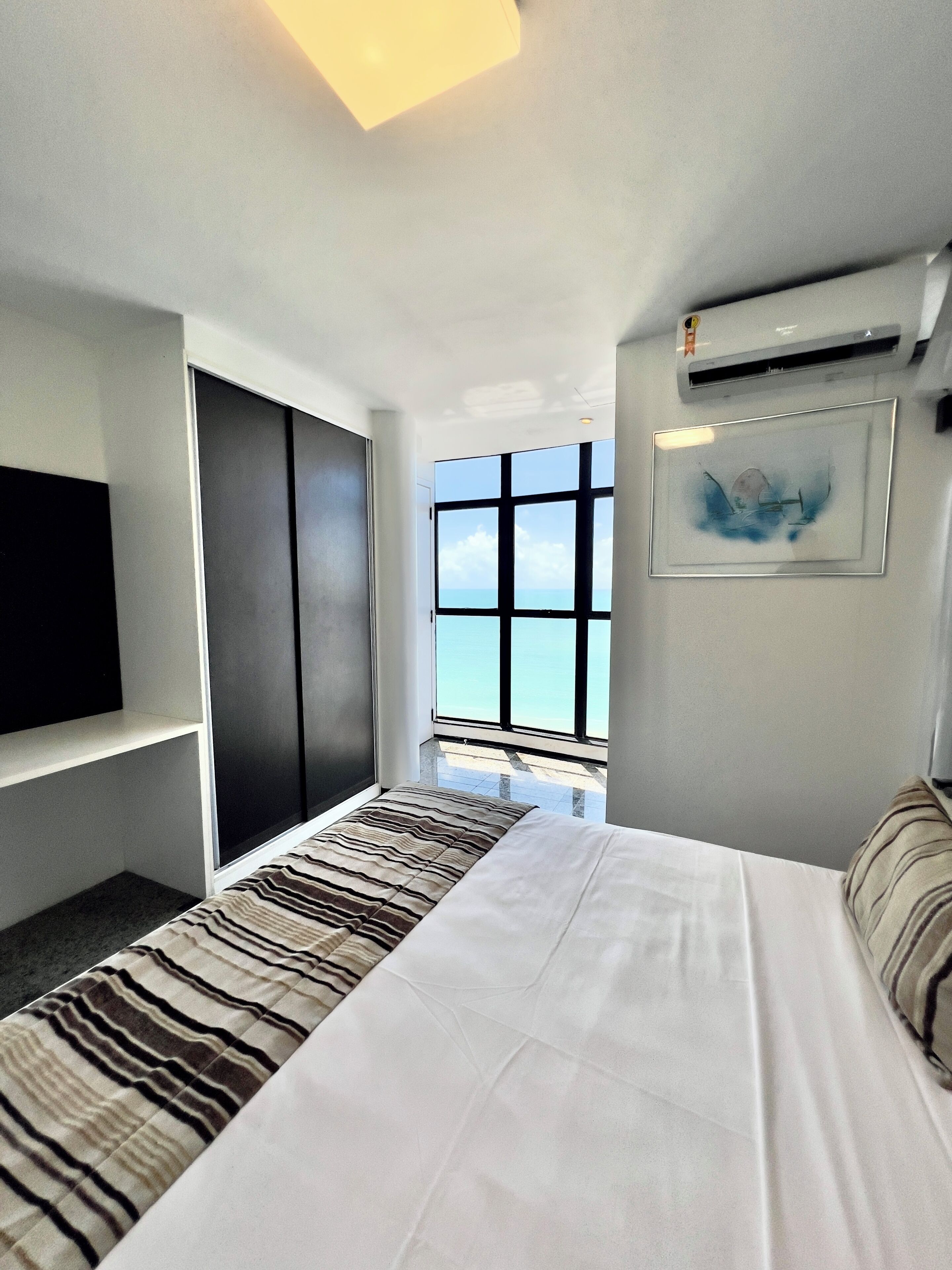 Quarto de hotel moderno com uma cama grande, televisão na parede, janela com vista para o mar e ar condicionado.