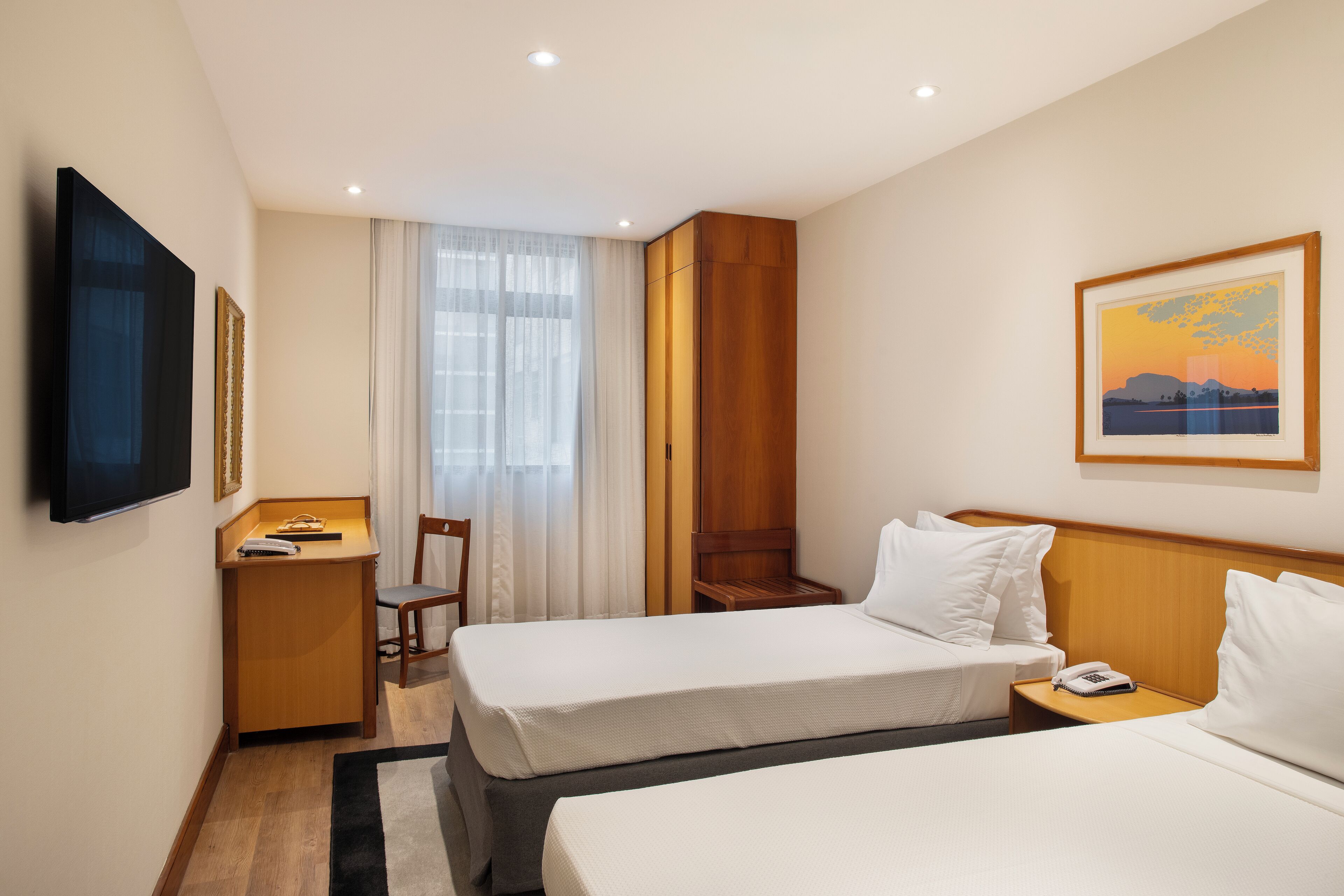A hotel room with two beds, a desk, a chair, a TV, and a painting on the wall.