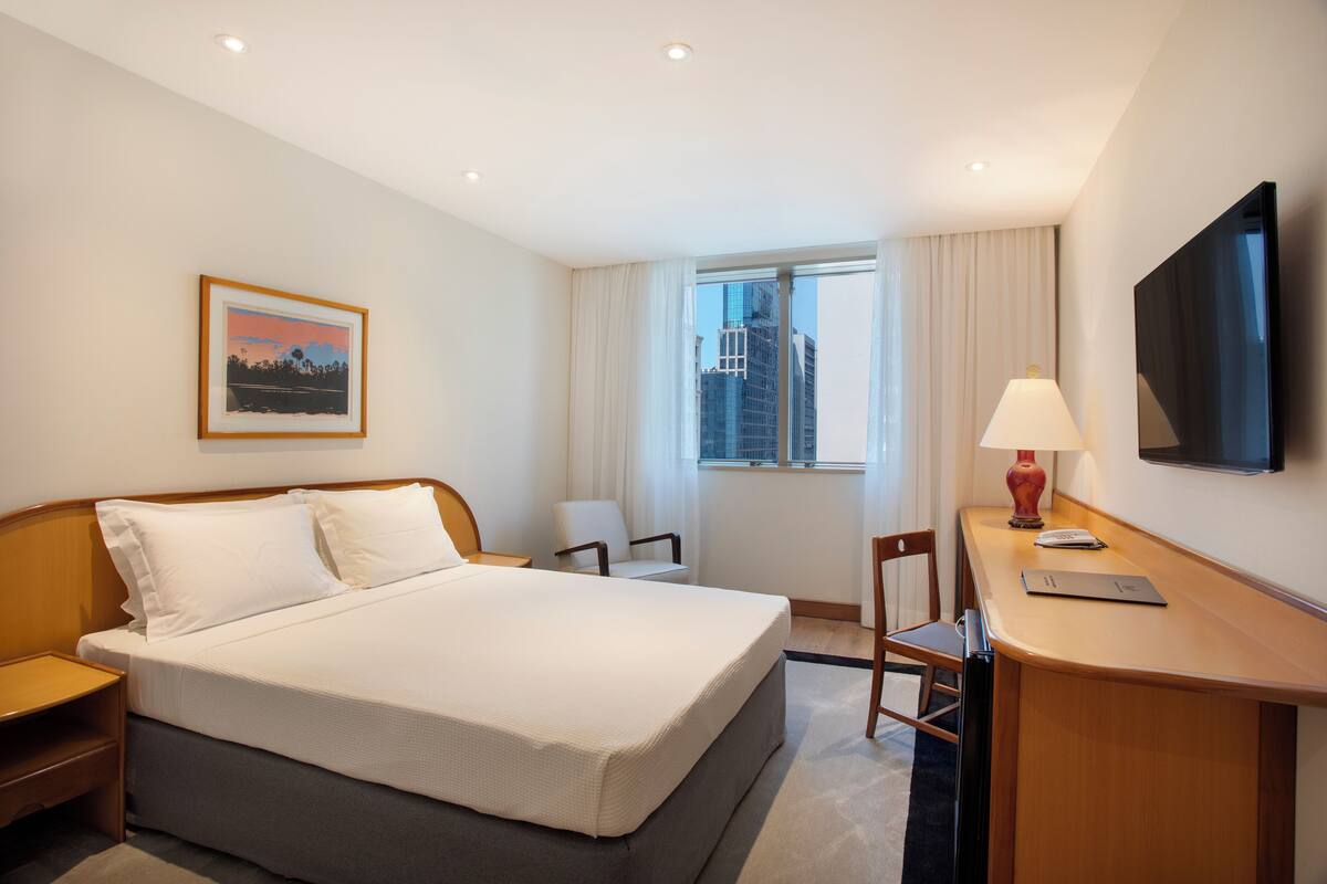 Hotelkamer met een bed, bureau, stoel, televisie, raam met gordijnen en een lijst aan de muur.