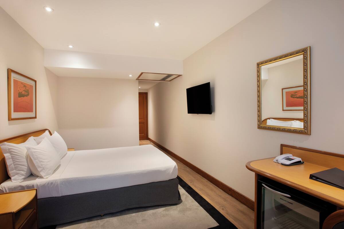 Een hotelkamer met een bed, nachtkastje, een bureau met telefoon, spiegel en een televisie aan de muur.
