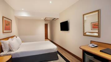 Una habitación de hotel con cama, mesita de noche, escritorio con teléfono, espejo y televisión en la pared.