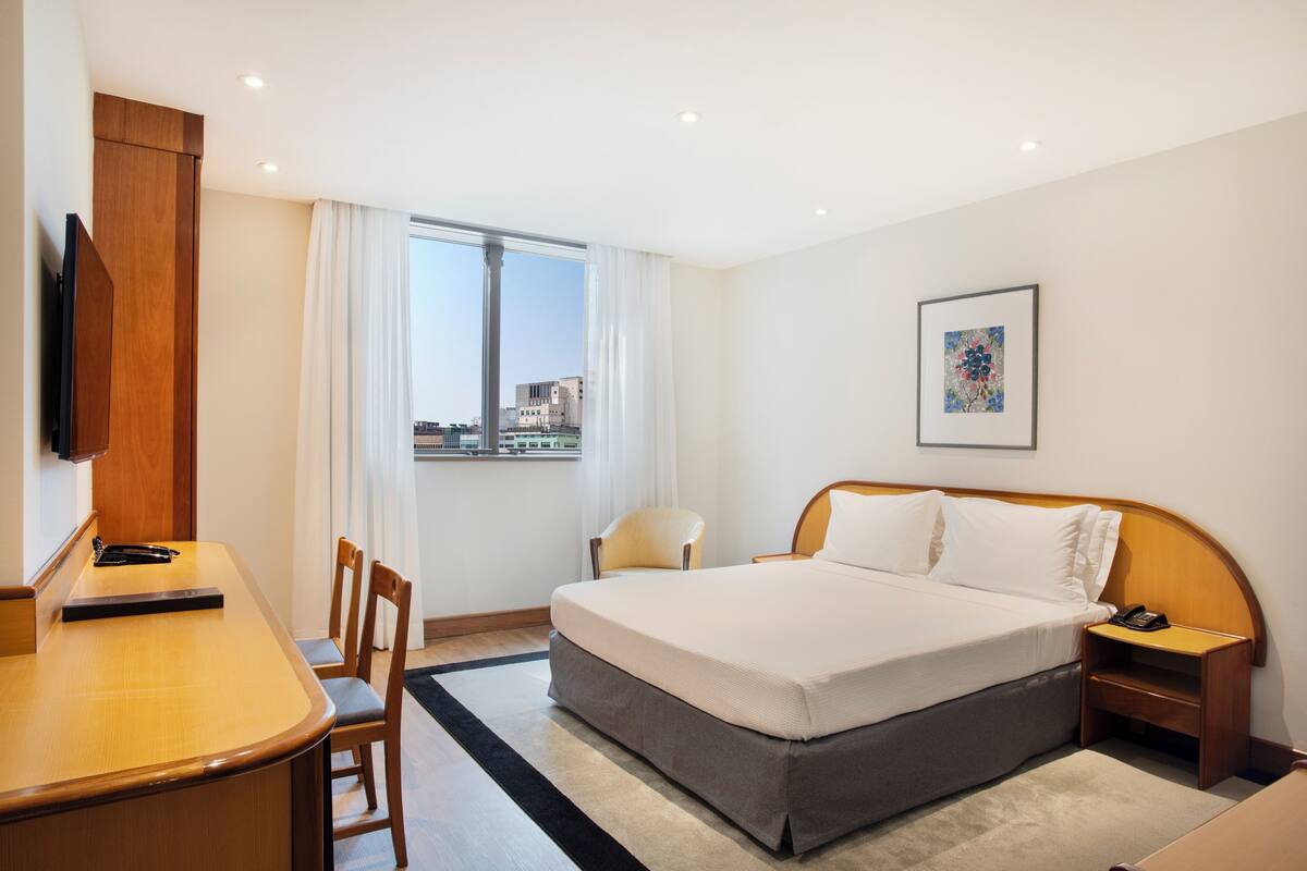 Hotelkamer met een bed, bureau, stoel, nachtkastje, televisie en een raam met uitzicht op de stad.