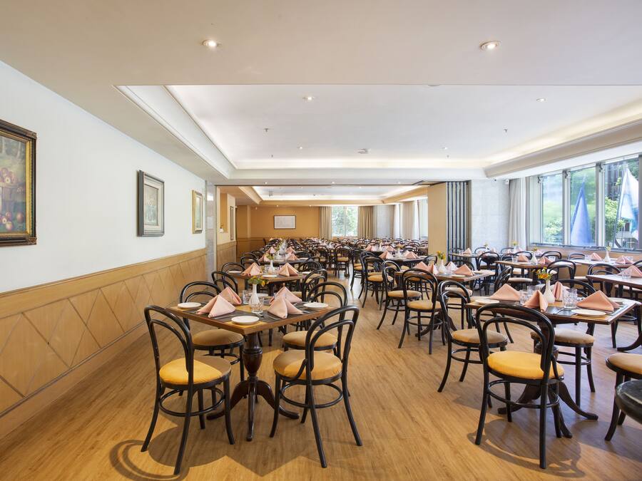 A spacious dining area with multiple tables and chairs, arranged in rows, with large windows providing natural light.
