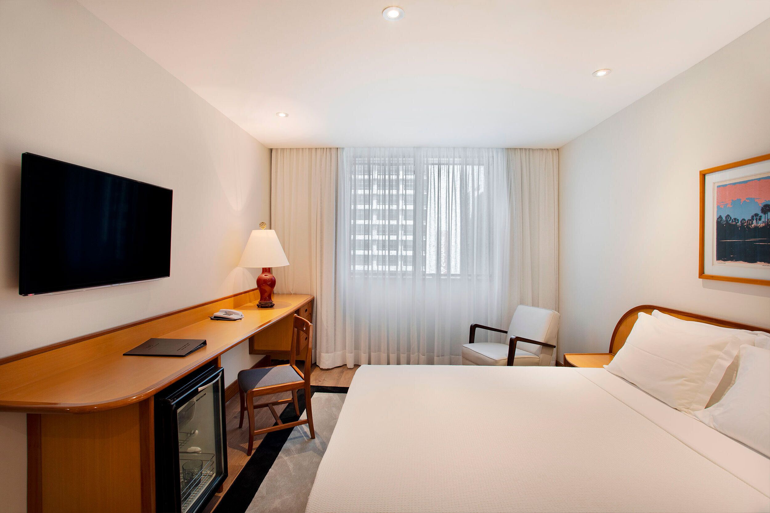 A hotel room with a bed, a desk with a computer, a TV, a chair, and a window with curtains.