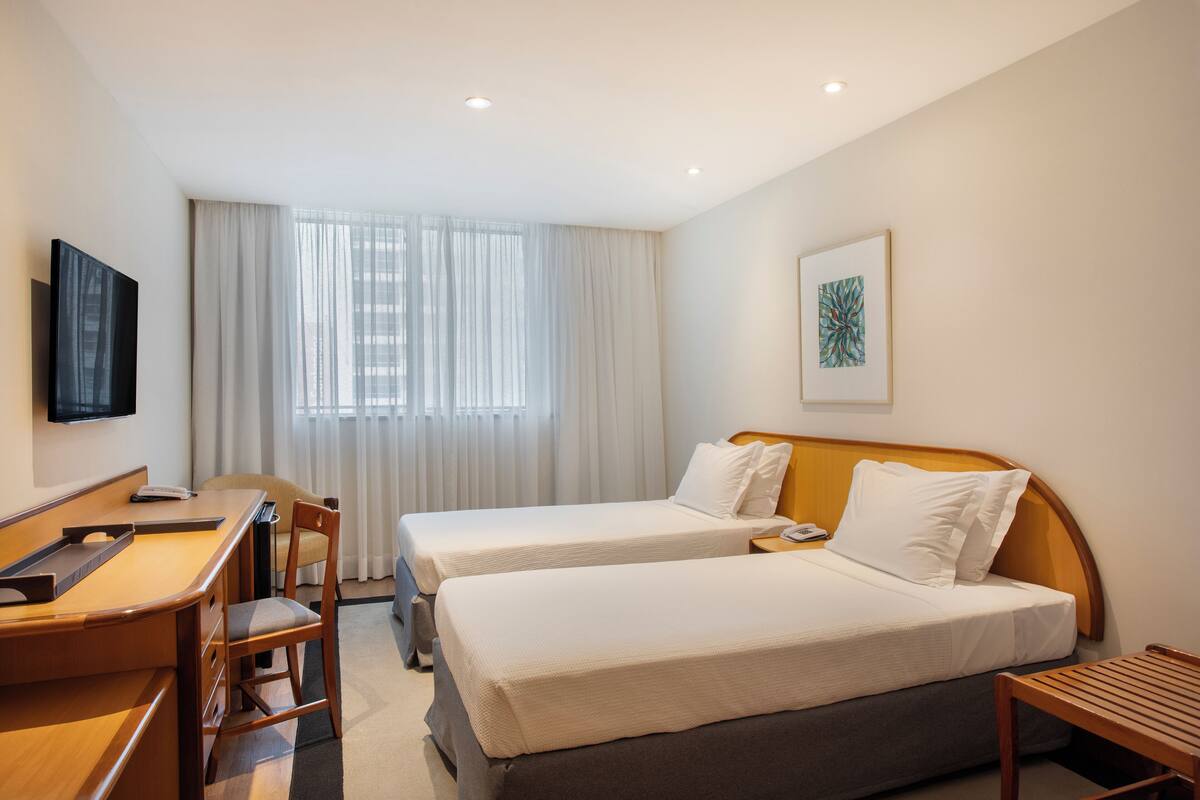 Een hotelkamer met twee eenpersoonsbedden, een bureau met stoel, een aan de muur bevestigde televisie en een raam met gordijnen.