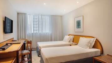 A hotel room with two single beds, a desk with a chair, a TV mounted on the wall, and a window with curtains.