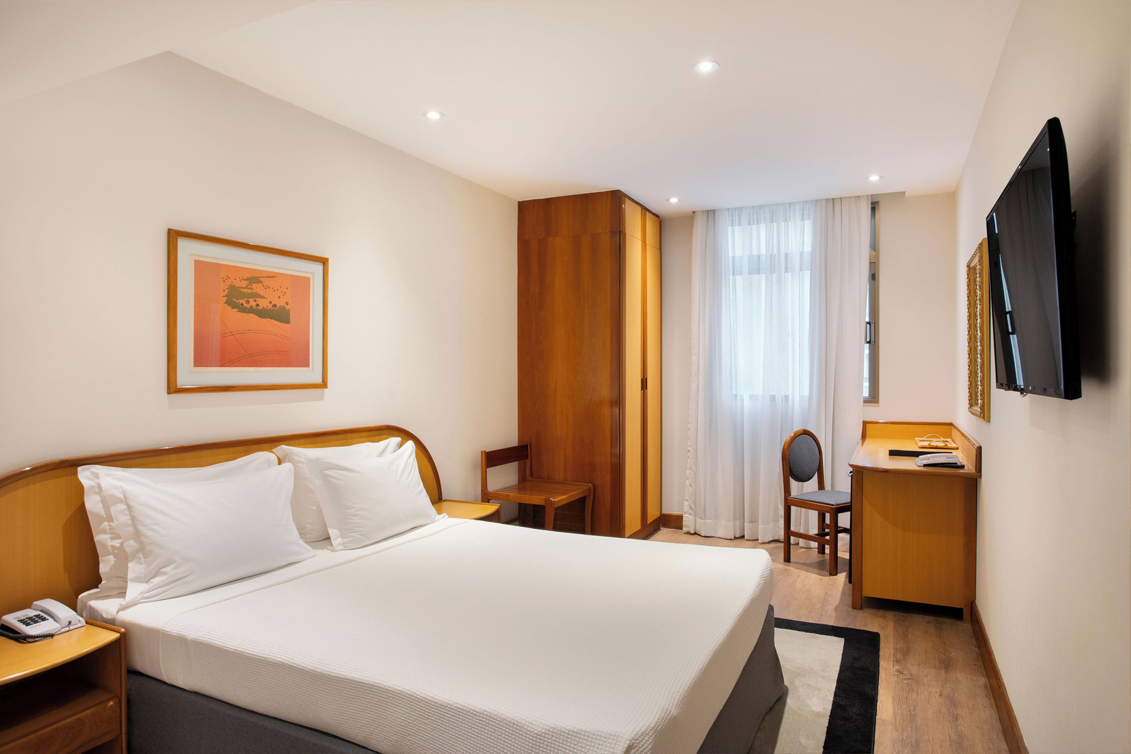 A hotel room with a bed, a desk, a chair, a telephone, a television, and a framed picture on the wall.