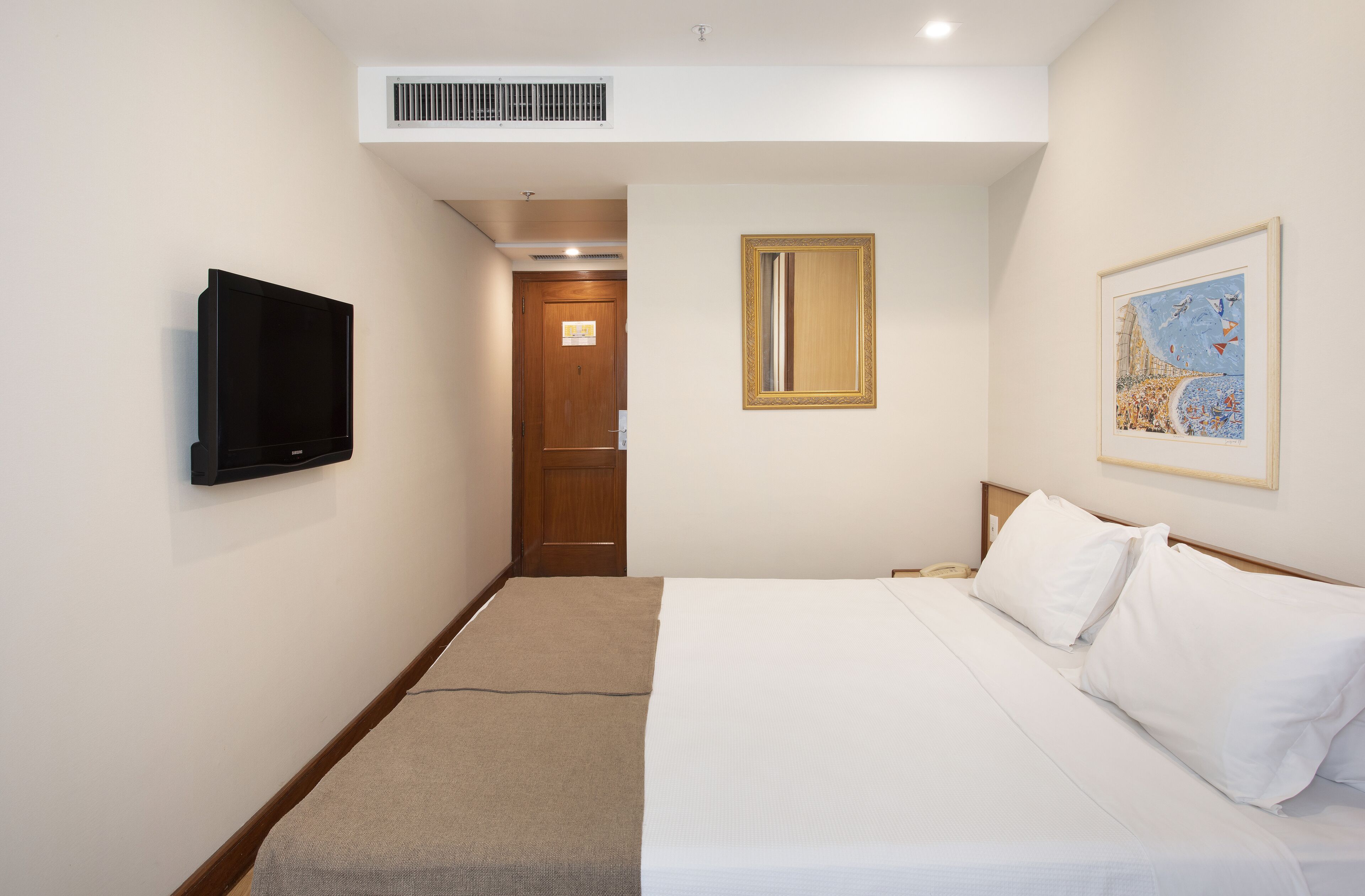 A hotel room with a bed, a television, a door, and a painting on the wall.