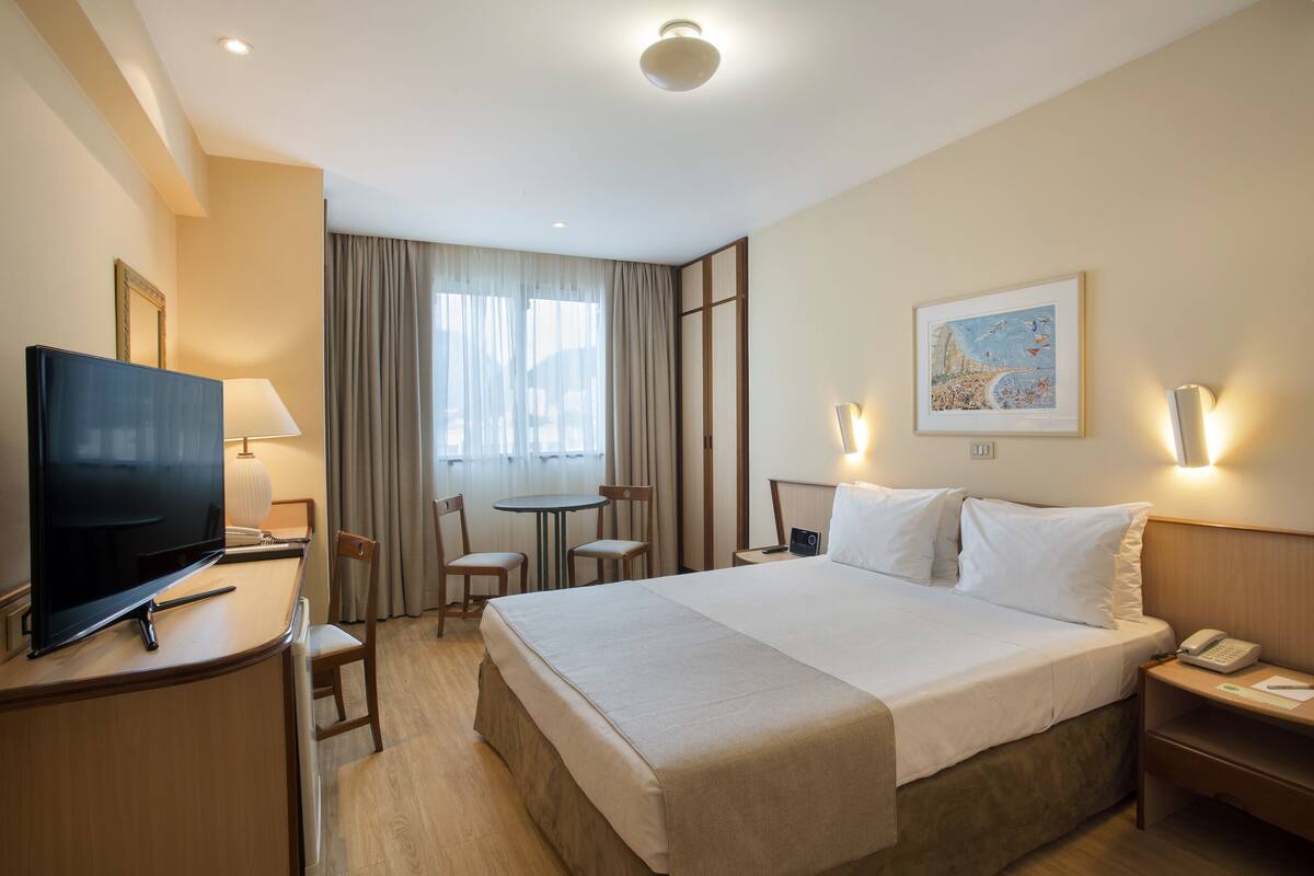 Une chambre d’hôtel avec un grand lit, un bureau avec une télévision, une chaise, une petite table et une table de chevet avec un téléphone.