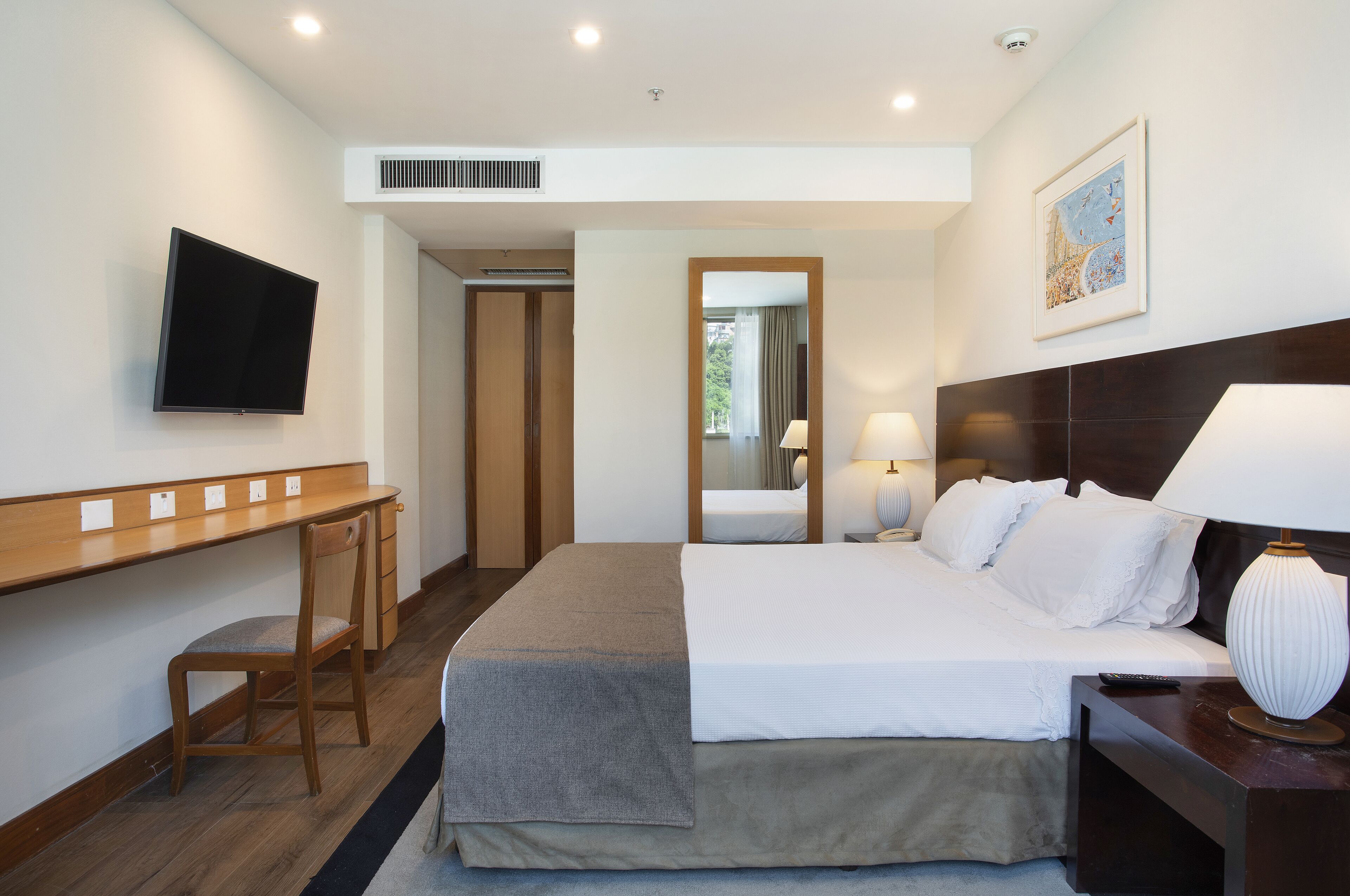 A hotel room with a bed, a desk, a chair, a TV, a lamp, and a painting on the wall.