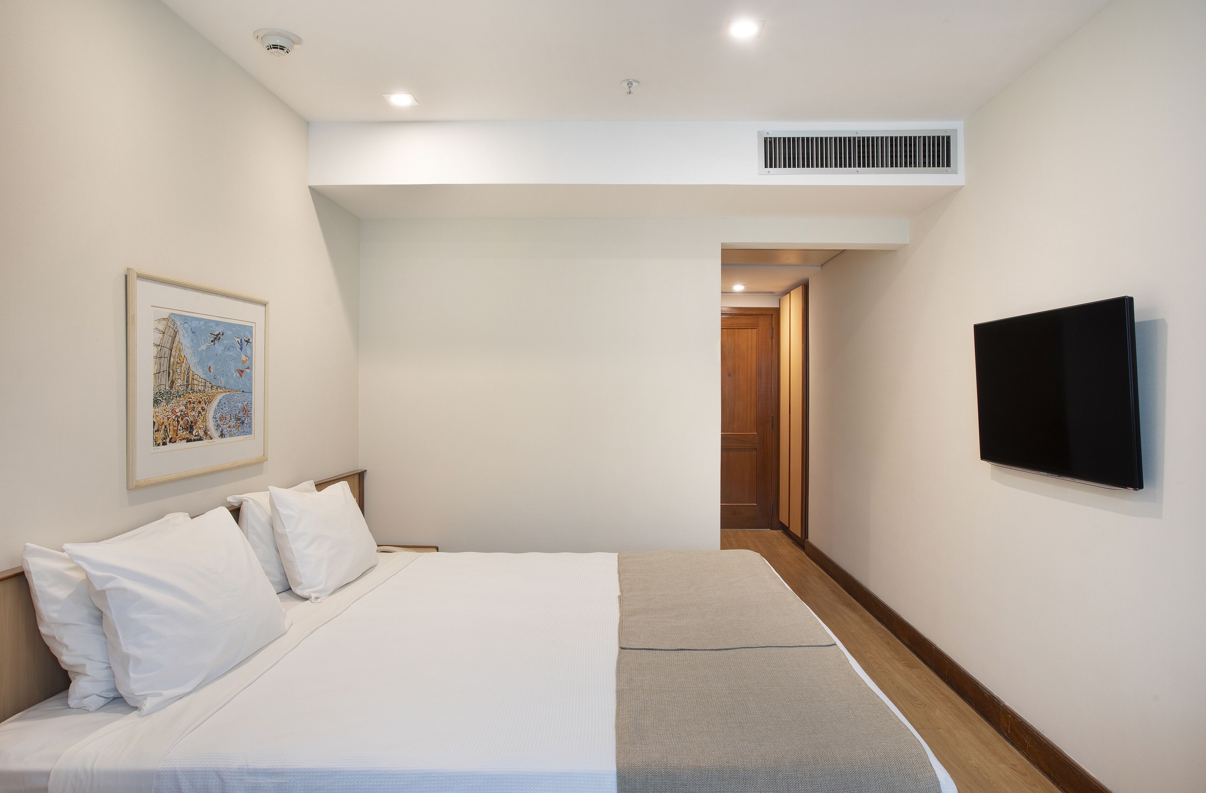 A hotel room with a bed, a flat-screen TV mounted on the wall, a framed picture on the wall, and a door leading to another room.