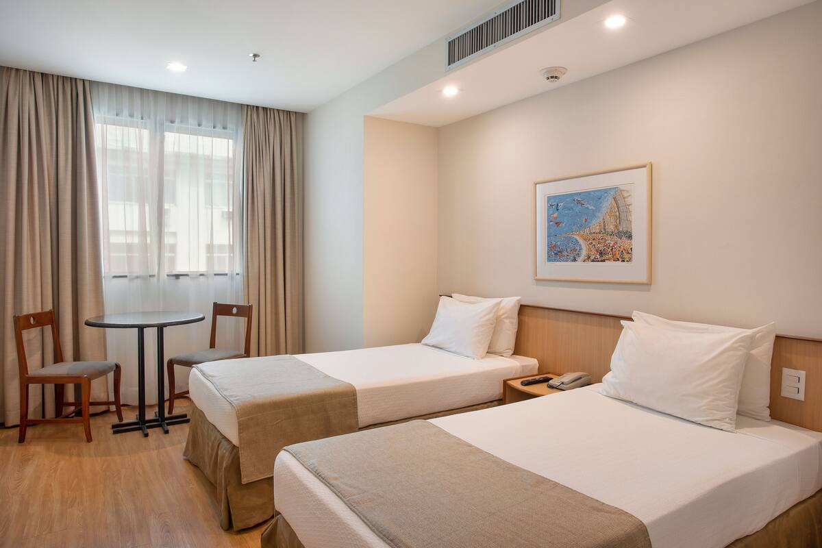 Une chambre d’hôtel avec deux lits, une petite table, deux chaises, un tableau au mur et un téléphone sur la table de chevet.