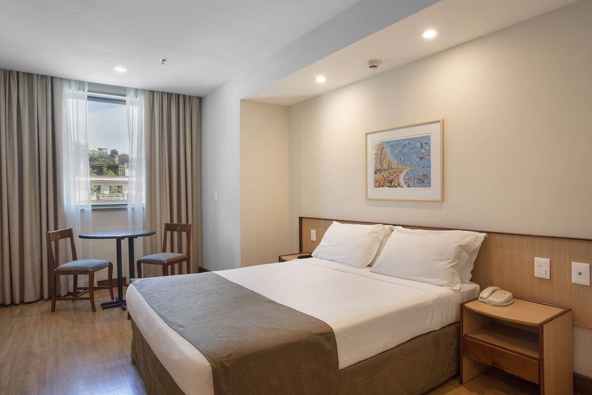 Une chambre d’hôtel comprenant un lit, une table de chevet avec un téléphone, une petite table avec des chaises et une fenêtre avec des rideaux.