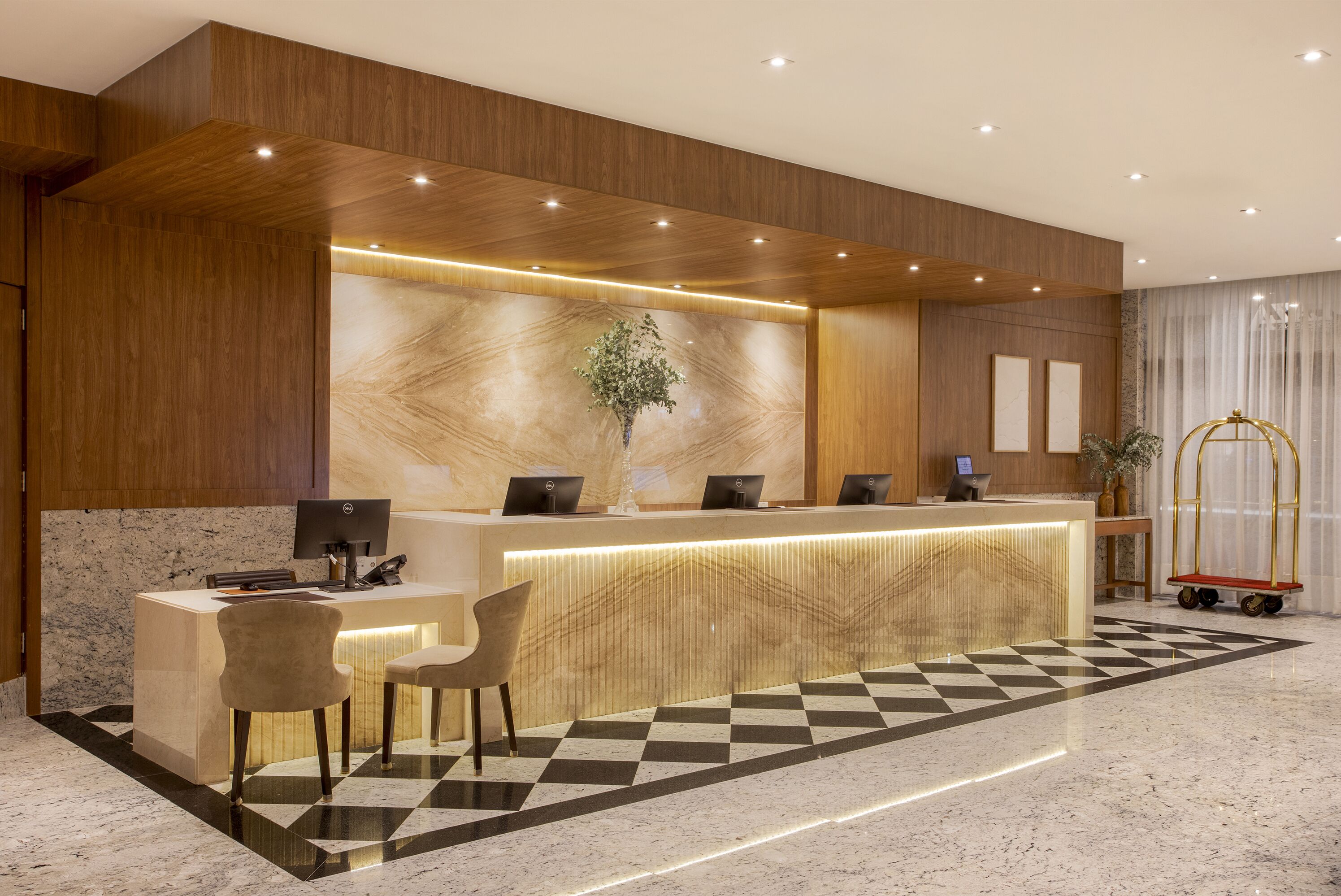 A hotel reception area with a wooden reception desk, two chairs, a plant, and a check-in counter.