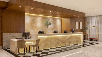 A hotel reception area with a wooden reception desk, two chairs, a plant, and a check-in counter.