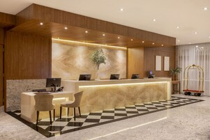 A hotel reception area with a wooden reception desk, two chairs, a plant, and a check-in counter.