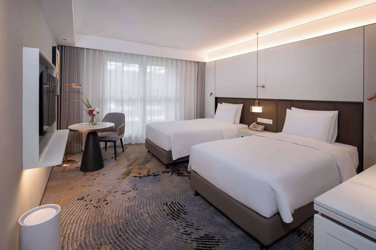 A hotel room with two beds, a desk with a chair, a TV, and a large window with curtains.