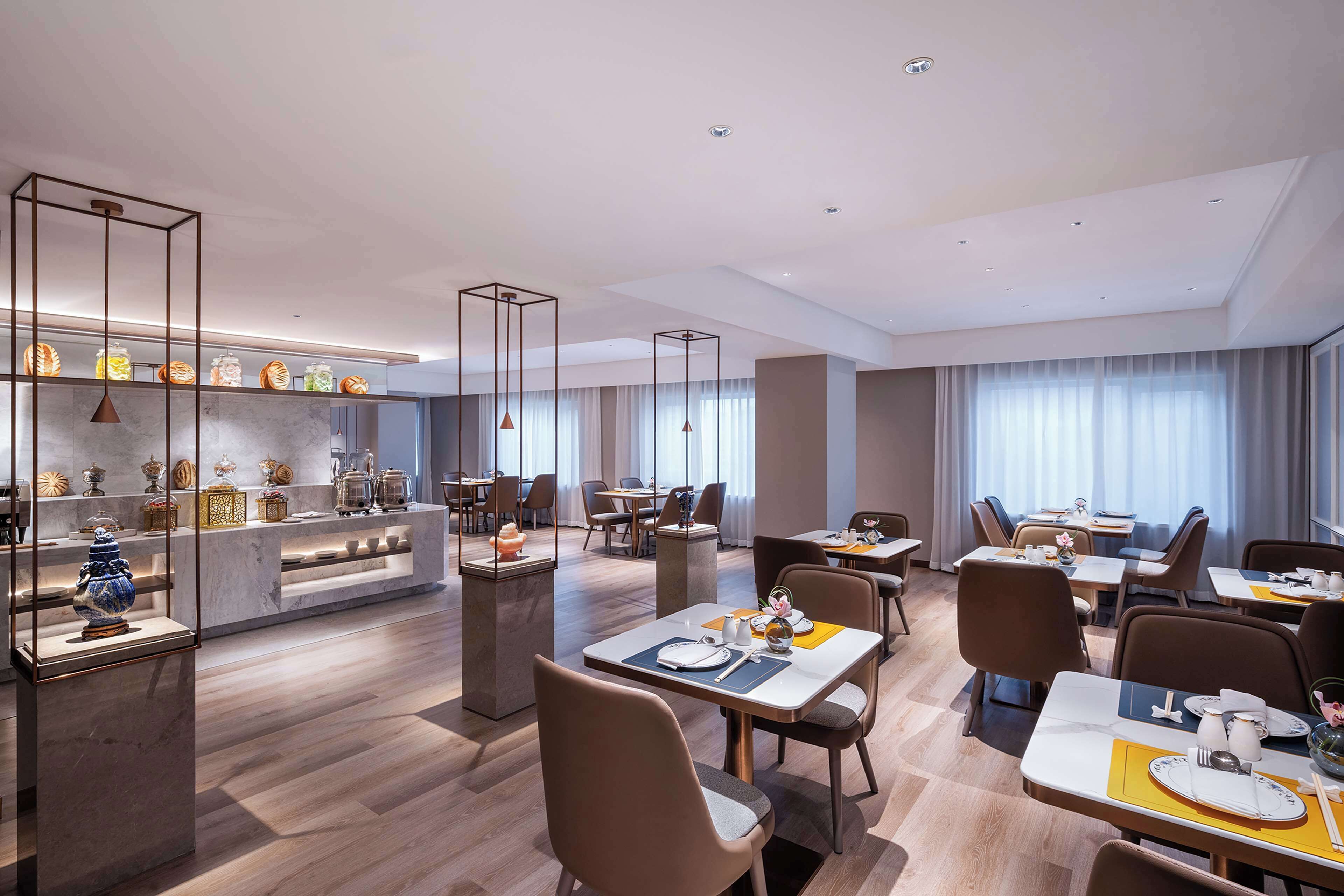 A modern dining area with tables and chairs, a display of decorative items on shelves, and a large window with curtains.