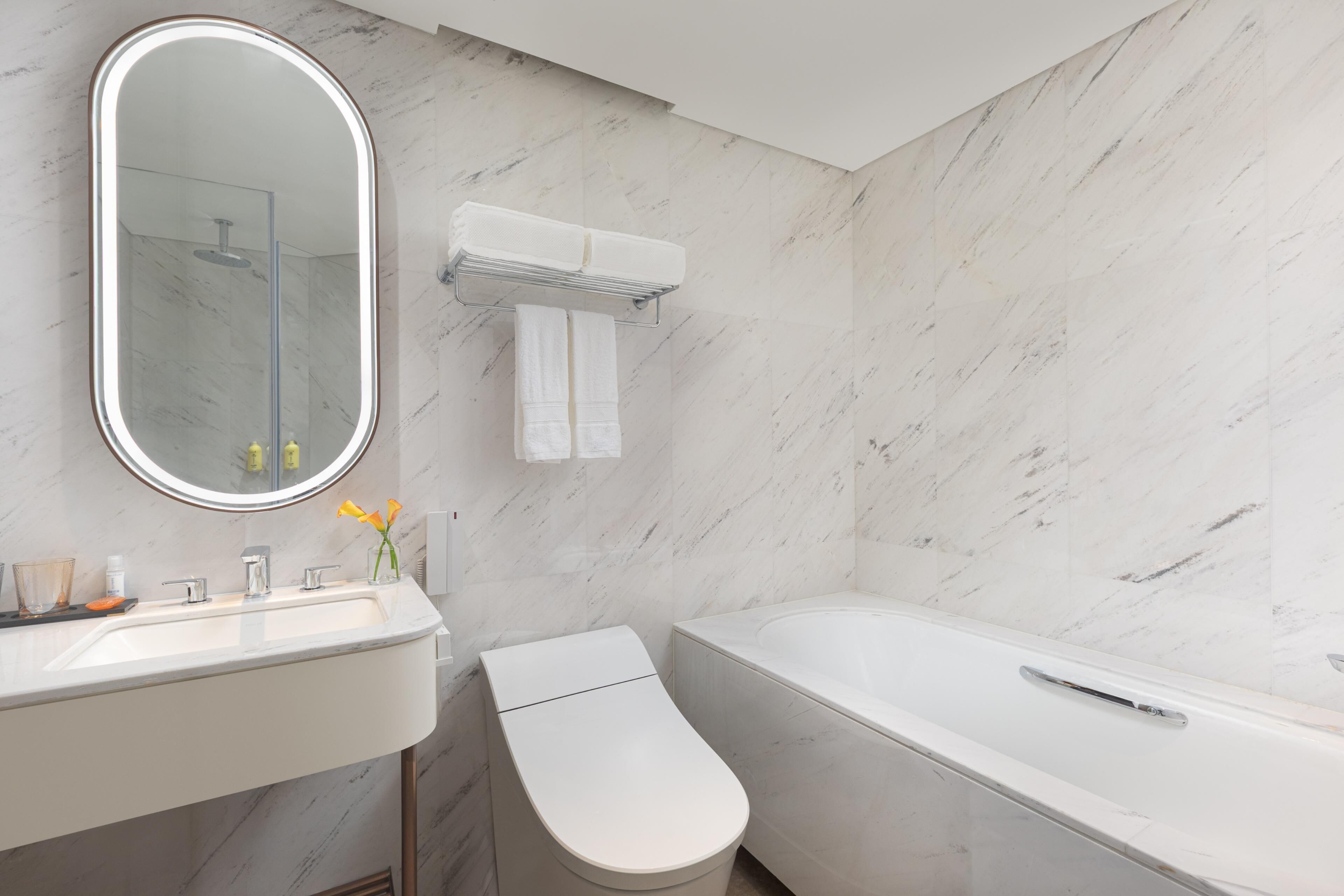 Un baño moderno con pared de mármol, un espejo redondo con luz, un lavabo blanco, inodoro y bañera.