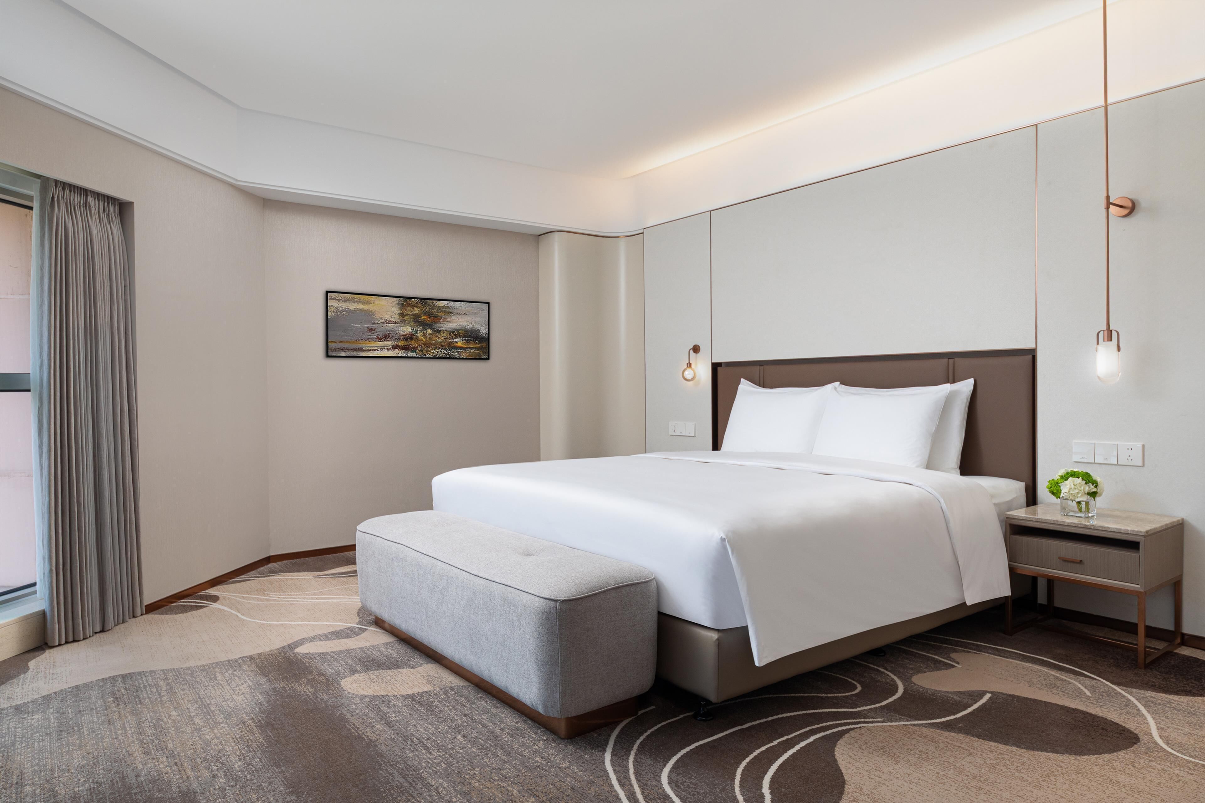 Hotellihuone, jossa on sänky, yöpöytä, lamppu, ikkuna verhoilla ja taulu seinällä.