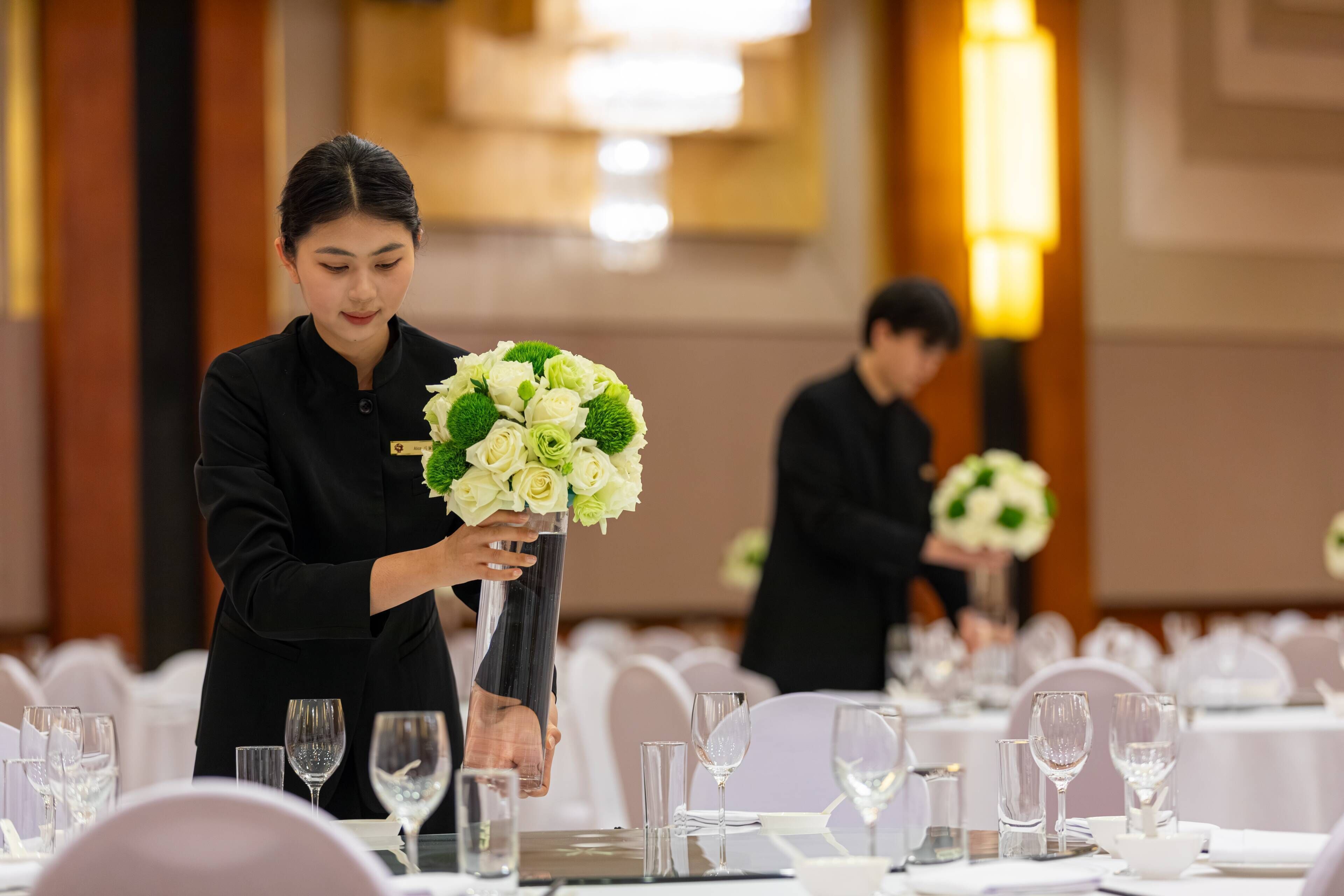 Une salle de banquet avec des tables dressées pour le repas, une personne qui arrange un centre de table floral, et une autre personne en arrière-plan.