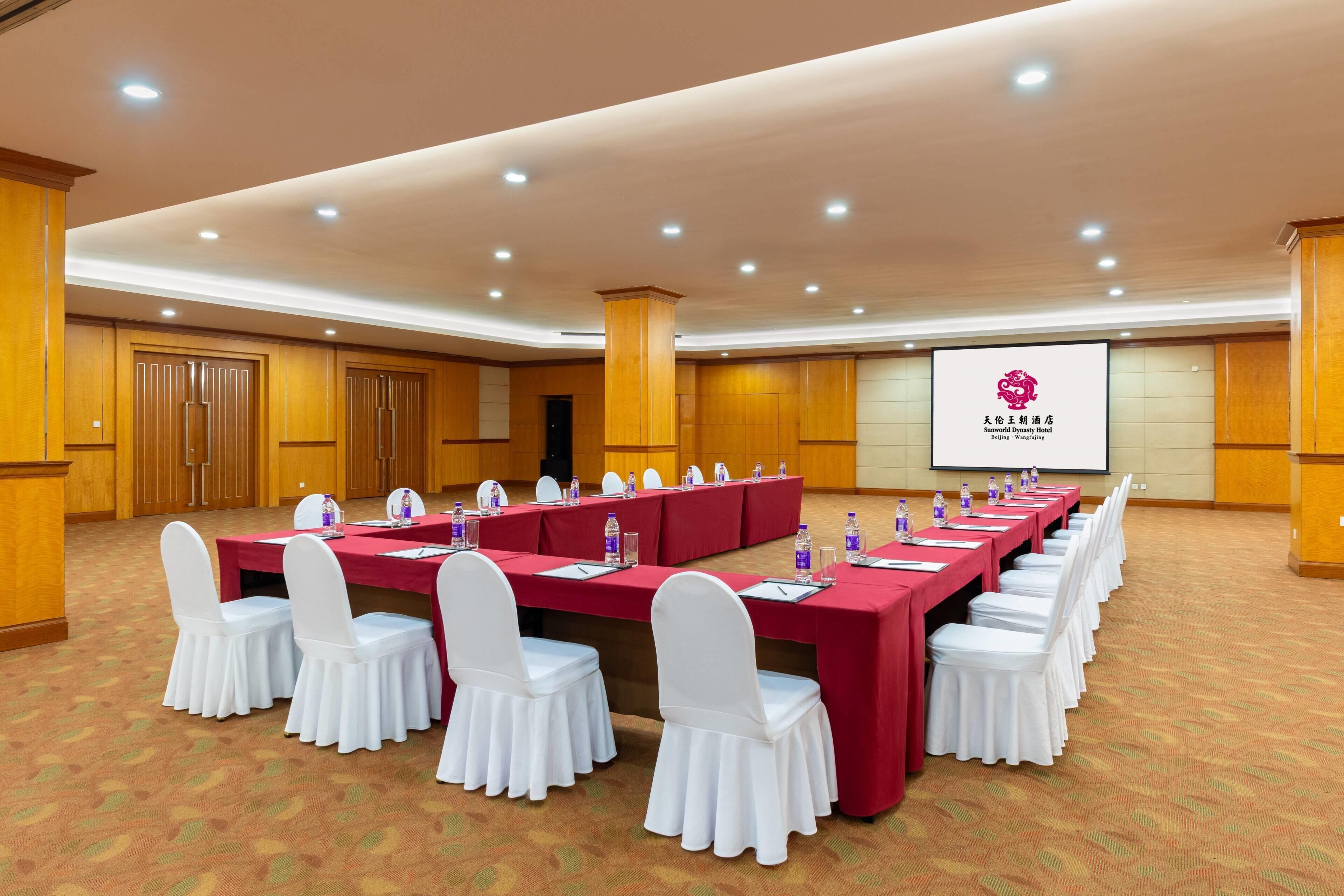 Een vergaderzaal met lange tafels, witte stoelen en een projectiescherm waarop het logo van Sunworld Dynasty Hotel staat.