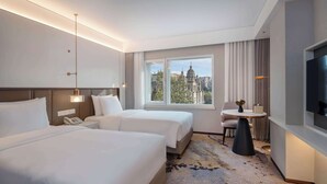 Ein Hotelzimmer mit zwei Betten, einem Fernseher, einem kleinen Tisch mit Lampe und einem Fenster mit Blick auf die Stadt.