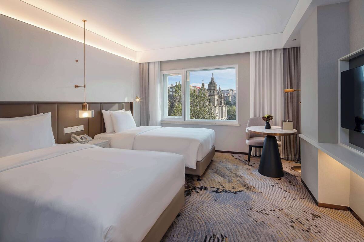 A hotel room with two beds, a TV, a small table with a lamp, and a window with a city view.