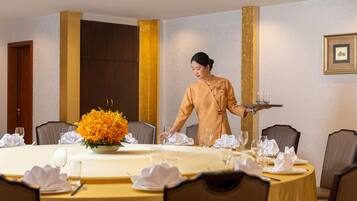 A hotel dining area with a round table set for a meal, a waiter carrying a tray, and a large floral centerpiece.