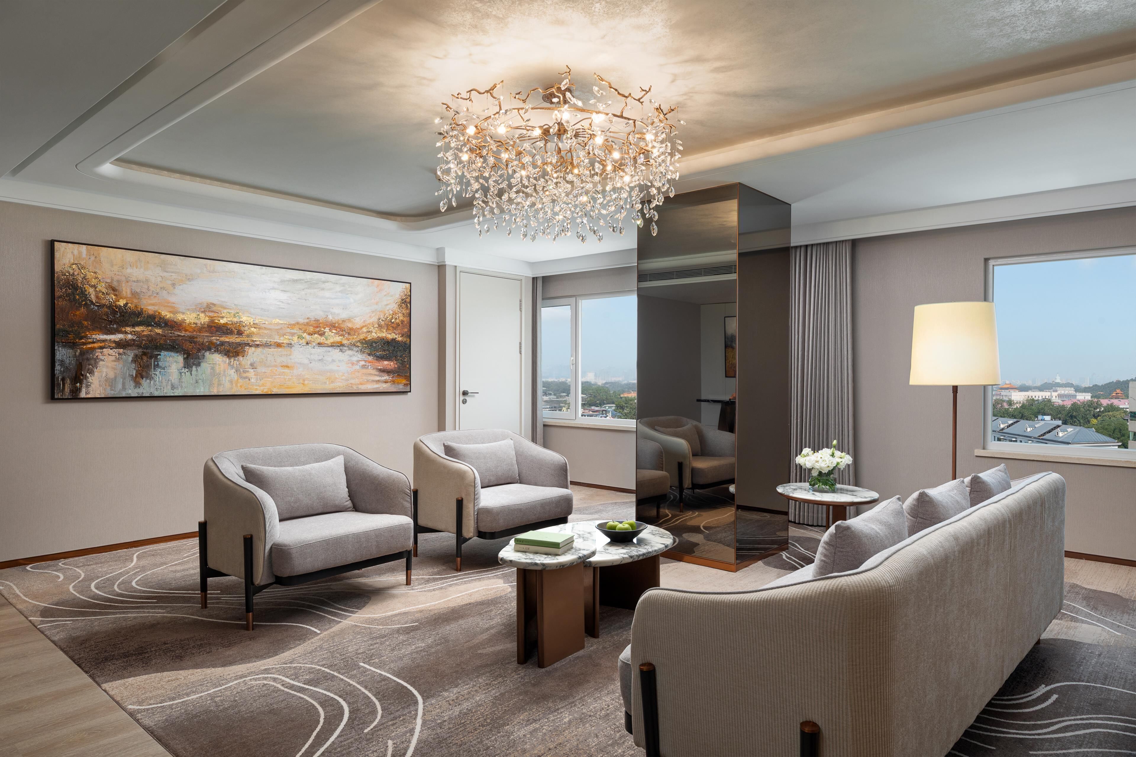 A modern hotel room with a large painting, a chandelier, two sofas, a coffee table, and a view through the window.