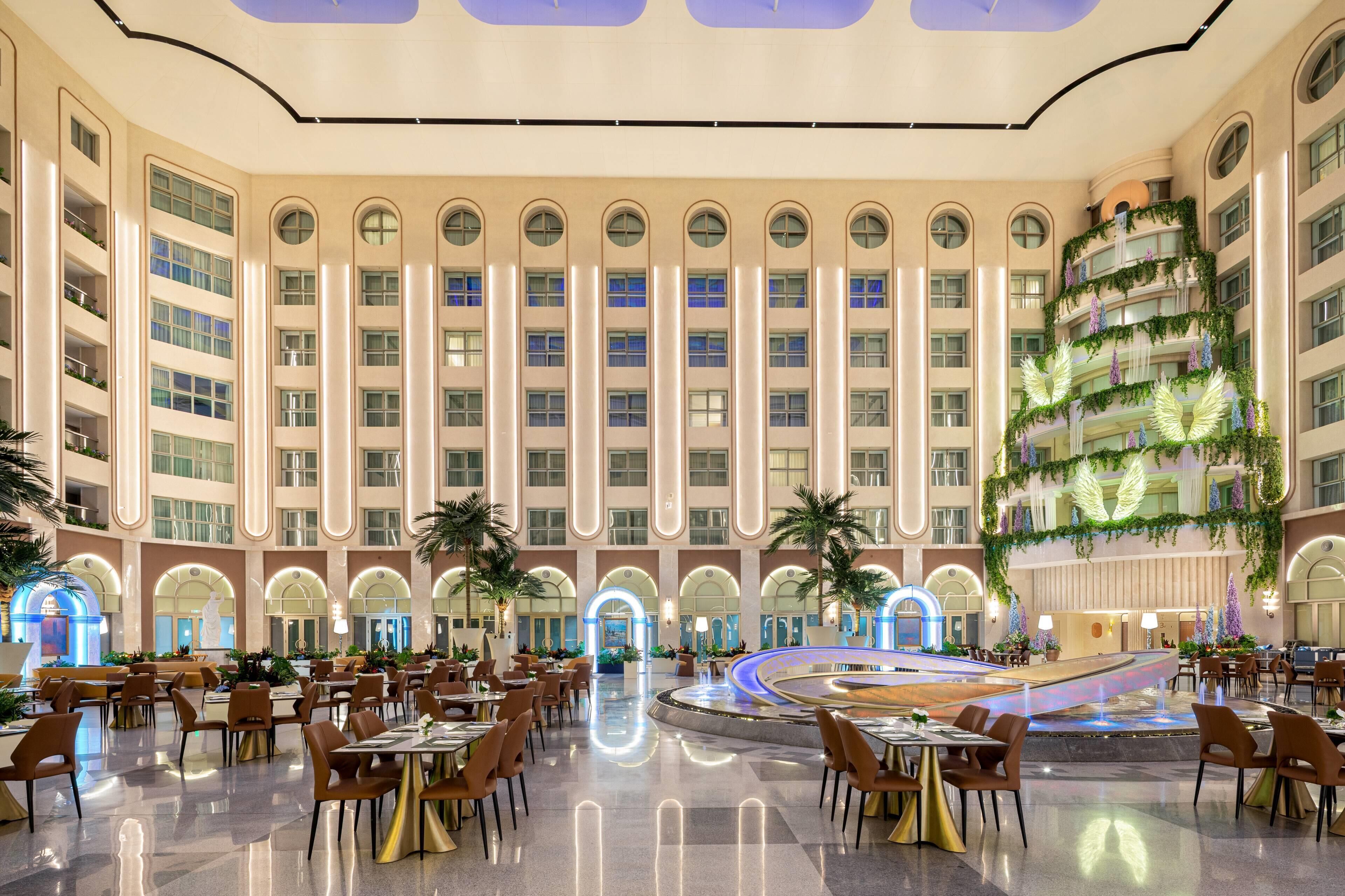 A spacious hotel lobby with a central fountain, multiple dining areas, and a large atrium with a decorative tree.